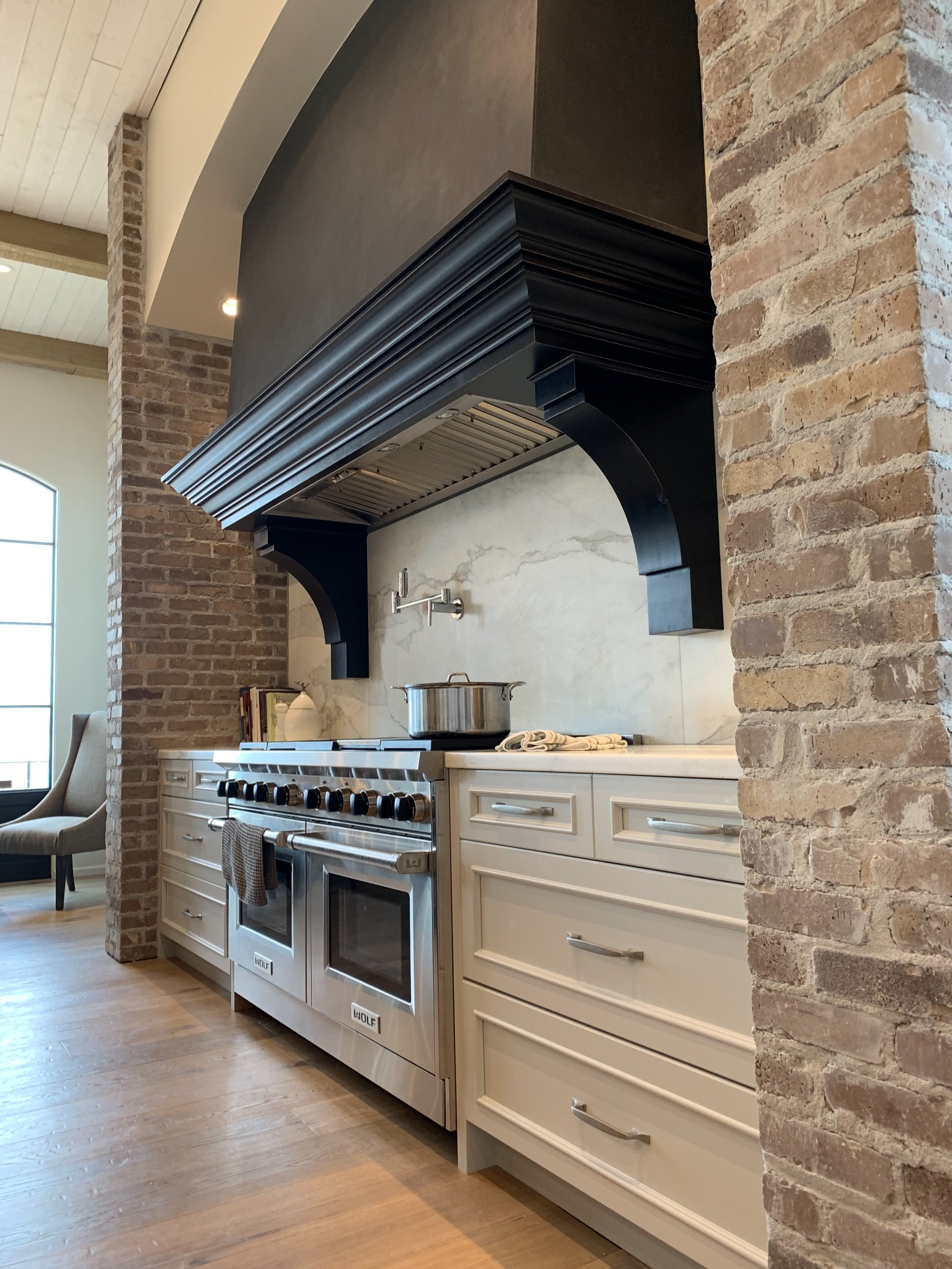 Black architectural range hood installed between brick columns above white cabinetry